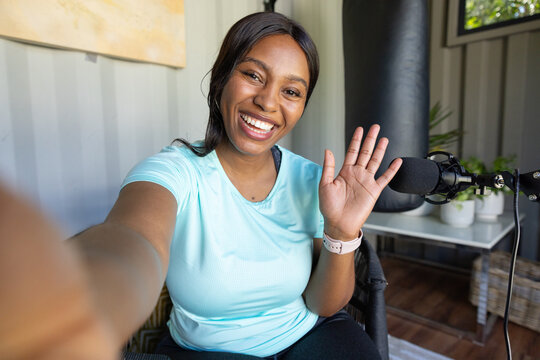 African woman sitting in home studio taking selfie while waving by mic, punching bag, smartwatch