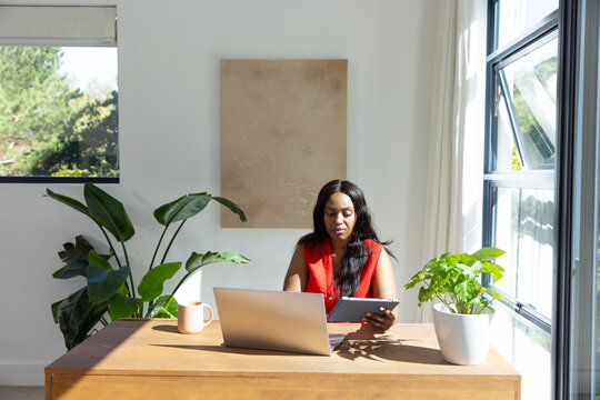 African woman wearing sleeveless red blouse working on tablet in home office with laptop plant mug