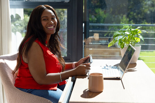 Woman sitting at wooden desk at home by balcony holding phone, laptop, plant, mug, smiling