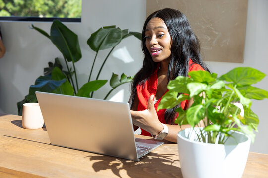 African American woman speaking gesturing working on laptop at wood table in red blouse amid plants