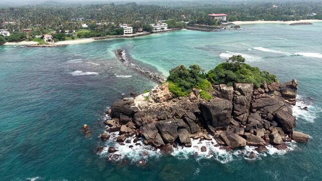 Aerial view of Devil's Rock islet with waves crashing against dark stones and a sandy beach in the distance, Sri Lanka.