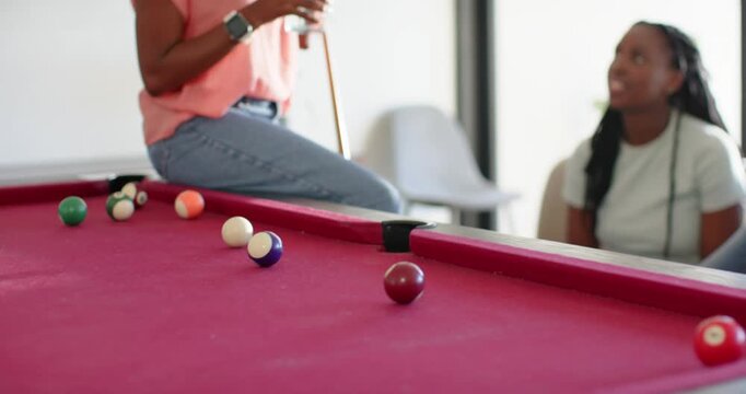 Camera pulling back revealing African American women holding cue and drink on pool table, chatting