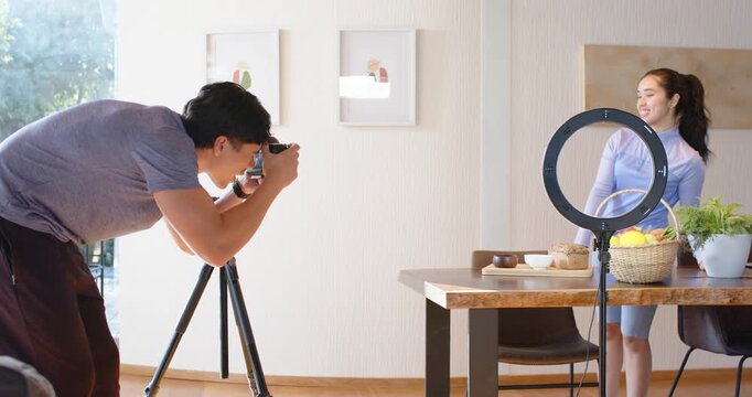Asian man leaning into camera shooting woman posing by dining table with ring light for content