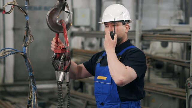 Industrial worker in hard hat using walkie talkie near crane hook in factory workshop. Technician coordinating lifting process in manufacturing facility.