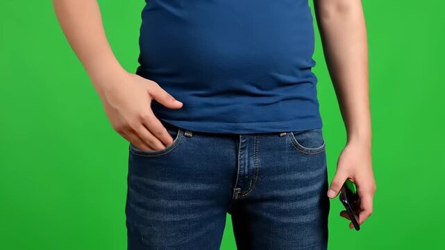 Person in blue t-shirt and jeans with hand in pocket, holding a smartphone, on a green screen background, close-up of midsection