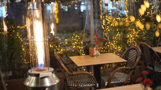Cozy restaurant terrace at night tall gas patio heater flame, empty wooden table wicker chairs surrounded by warm fairy lights, plants clear plastic wind screens, romantic outdoor dining, no people