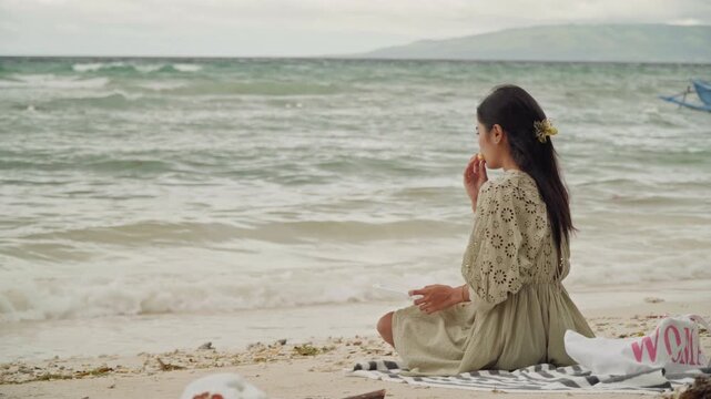 Asian woman meditative beachside sitting on blanket facing gentle ocean, vintage dress and floral hair clip, shells and driftwood on sand, calm waves, overcast sky, reflective mood, cinematic slow