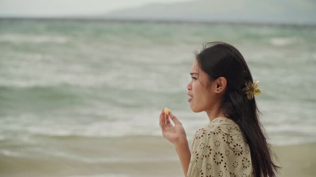 Asian woman beachside profile tasting snack by rolling waves, floral hair clip and patterned dress, small tropical bite, pensive expression and distant gaze, candid travel portrait, seaside taster