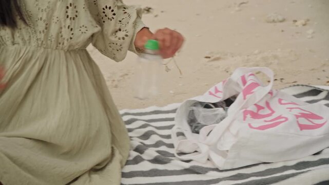 Woman on beach towel preparing picnic, unpacks pink tote, smooths striped blanket, reaches for capped water bottle, removes lid and sets cup, boho eyelet dress fluttering, sunlit sand and calm seaside