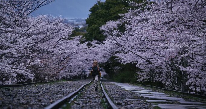 Happy, funny young boy with backpack running through gravel on rails with surrounding cherry blossom trees in slow motion