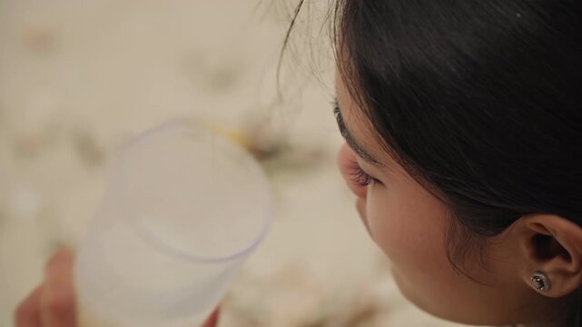 Young woman sipping drink on beach, closeup of plastic cup and sandy hands, profile shots reveal pensive gaze, scattered leaves and shells on shoreline, warm afternoon light and tranquil mood