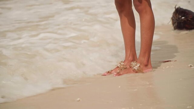 Bare feet on wet sand, shoreline. Closeup of female legs wearing embellished sandals stepping through shallow waves, soft foam lapping at ankles, warm golden light, sandy footprints left behind,