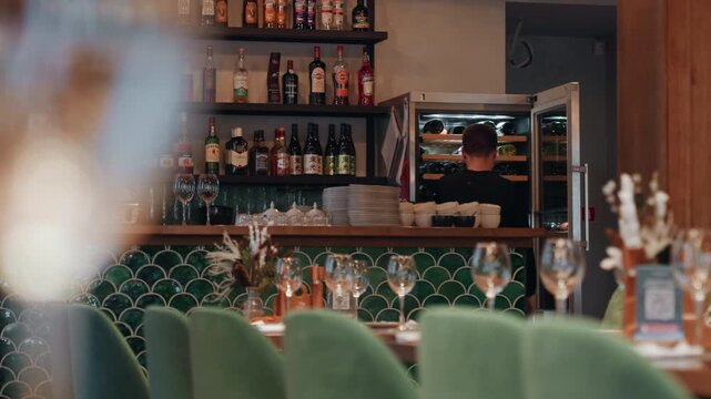 Cozy restaurant bar green chairs showing velvet seating and set tables, sparkling glassware and floral accents, bartender arranging bottles on shelf and opening fridge, server polishing stemware,