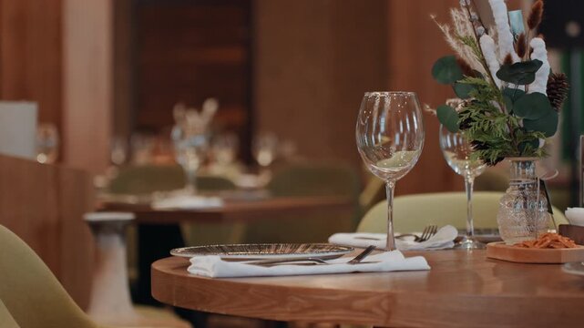 Intimate restaurant table setting warm floral glassware and cutlery arranged on wooden round table, vase with dried flowers, soft bokeh background, empty green chairs, fine dining mood, server