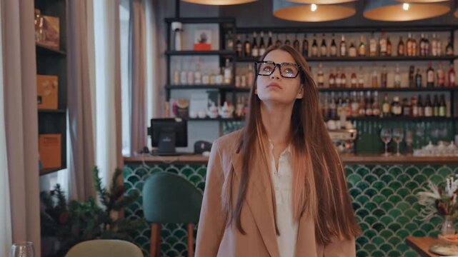 Young woman in cozy cafe bar, long brown hair and black glasses, wearing camel coat, standing near green tiled counter with bottlefilled shelves, soft warm lighting, empty tables set for brunch,