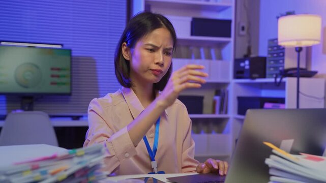 Stressed young Asian office worker feeling overwhelmed with paperwork and workload while working on laptop in office