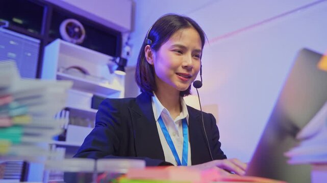 Smiling young Asian customer service representative wearing microphone headset and working on laptop in modern office.
