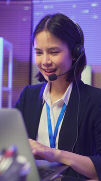 Smiling young Asian customer service representative wearing microphone headset and working on laptop in modern office.