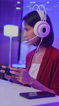 Excited young Asian girl playing video game with gaming headset and controller at home, esports online concept.
