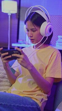Excited young Asian girl playing video game with gaming headset and controller at home, esports online concept.