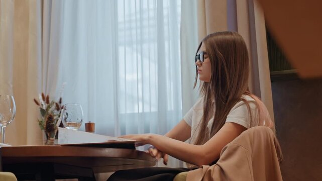 Caucasian woman rubbing forehead, freelance designer stressed over deadline, laptop and notes nearby, empty cup and glass on table, tense posture and anxious gaze toward window, dim warm light, urban