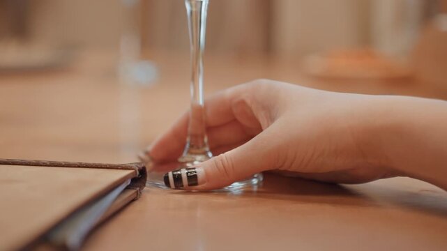 White woman adjusting wine glass base with manicured fingers, closeup on stem and tabletop, polished nails and ring detail, refined dining gesture captured in warm ambient light, elegant hospitality