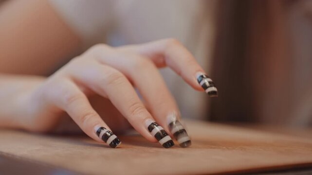 Closeup hand with striped black nails resting on wood table, beauty influencer in studio, warm cinematic lighting, glossy polish, delicate finger movement, refined nail design detail.