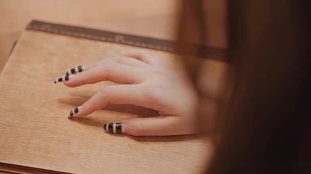 Woman writer hand on leather journal, closeup of manicured nails with black stripe polish, ruler accent, warm wood desk, soft ambient lighting, contemplative mood, creative planning gesture