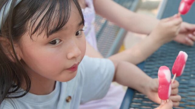 Two Young Asian Girls Enjoying Popsicles in a Sunny Outdoor Park