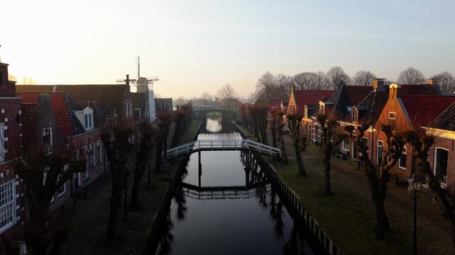Aerial view of the historic canal lined with traditional gabled houses and a windmill in the distance during golden hour in Sloten, Friesland, Netherlands.