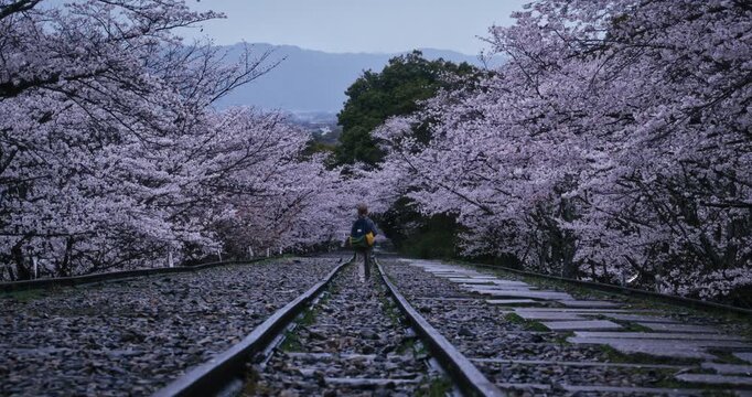 Little boy with backpack walking away on gravel train tracks in Japan then turning around - wide shot