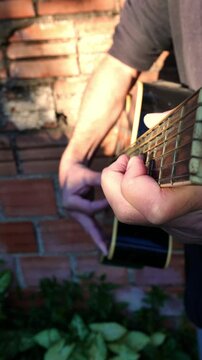 Slow zoom in: man practicing guitar. Musician plucking a guitar. Close-up of a man strumming an acoustic guitar under the sunlight. POV of a guitar neck. A person playing guitar outdoors. Close-up 