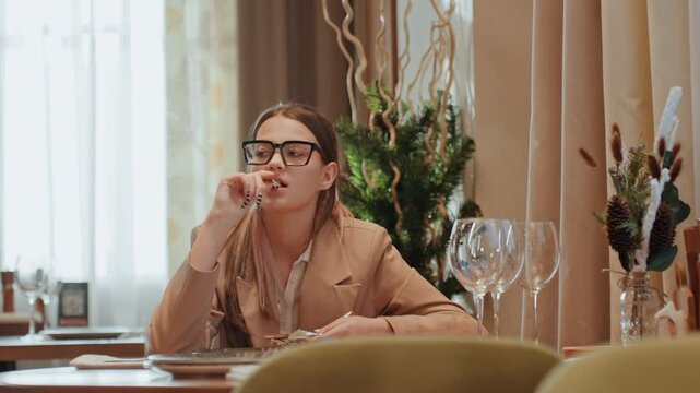 Businesswoman in cafe sampling dessert, checking notes and menu, wearing glasses, glassware and festive greenery on table, editorial vibe, contemplative expression, modern interior, quiet solo moment