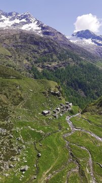 Aerial view of the historic stone houses of Alagna Valsesia nestled in a green valley with a stream and snow-capped mountains Alagna Valsesia, Piedmont, Italy.