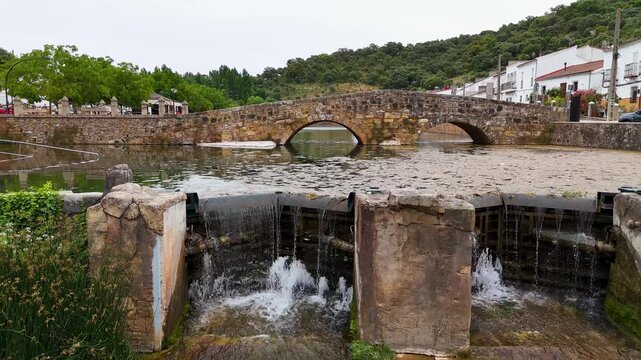 Aerial view of Puente de Piedra stone bridge over the Galapagar river with a small waterfall and lush greenery in San Nicolas del Puerto, Andalucia, Spain.