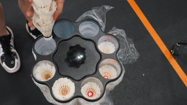Top down view badminton shuttlecocks loaded into circular feeder on indoor court, hands placing stacks of shuttlecocks, sneakers and orange boundary line visible, bubble wrap around holder, closeup