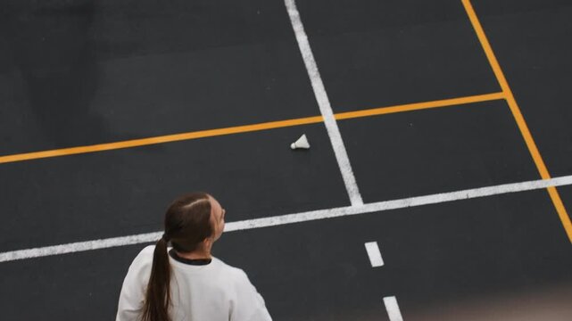 Focused badminton serve practice solo woman overhead court shot shows player preparing toss, steady stance, ponytail swing, shuttlecock on court, intentional rhythm, indoor gym lighting, concentrated