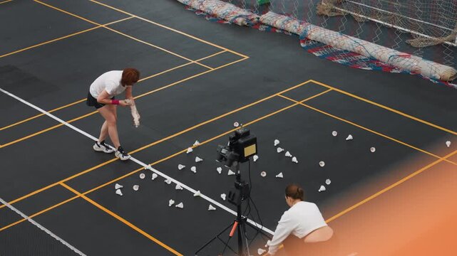 Player collects scattered shuttlecocks across court, quick crouch and reach movements, tripod camera records cleanup after highvolume rally drill, energetic atmosphere with focused routine
