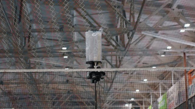 Automated shuttle machine launching multiple shuttlecocks over net, tripod mounted launcher in indoor gym, precise timed feed for player drills, metal structure and ceiling lights, equipment focused