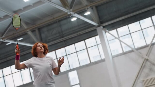 Caucasian woman practicing badminton in gym, executing overhead serves and smashes, neon racket and pink wristband visible, white shirt and shorts, high ceiling sports hall with large windows, solo