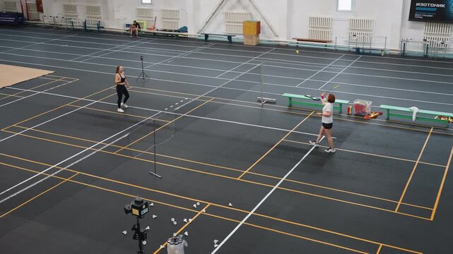 Indoor badminton training on court showcasing two players in fast paced rally, focused footwork and net exchanges, shuttlecock flight, marked lanes and benches, gym lighting and camera capturing