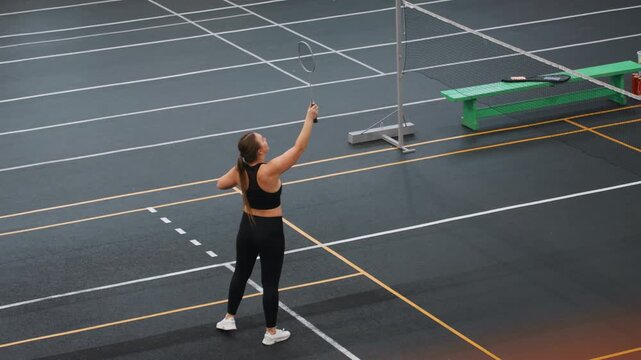 Indoor badminton court caucasian woman serving and retrieving shuttlecock, concentrated solo drill with precise toss and swing, repeated pickups and footwork adjustments, intensity and technique