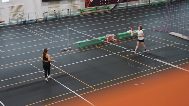 Indoor badminton court with caucasian women rallying, two players exchanging shuttlecock across net, light training drill and footwork practice, partner sparring and cooperative coaching moments, gym