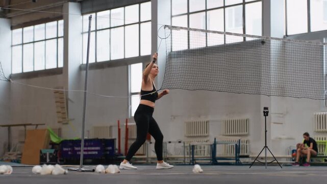 Sporty woman preparing for shuttlecock rally. Athletic woman in black attire warming up with shuttlecock drills. Female athlete in vibrant gym demonstrating precise serve and footwork techniques