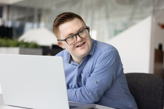 Young professional man with Down syndrome working on laptop seated at desk in modern corporate office, looks away with smile, feel satisfied with workflow results, engaged in remote job at workplace