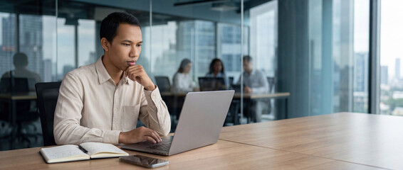 Obraz premium Busy professional 28-year-old Indonesian business man company employee, young male businessman looking at computer thinking on online investment or software technology using laptop working at office. 