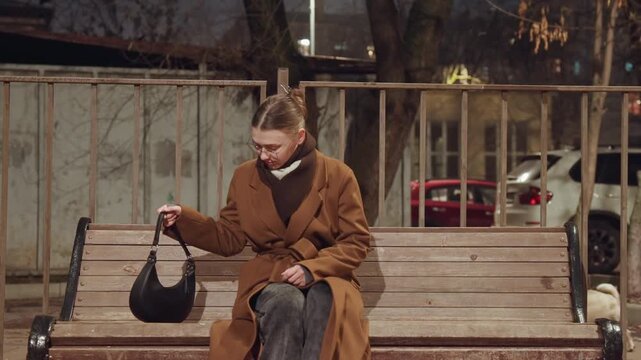 Caucasian woman sitting alone on bench in evening, brown coat and glasses placing handbag beside, contemplative posture with urban fence and parked cars backdrop, calm reflective pause during commute