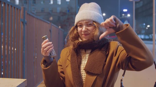 White woman inspects bottle at night, evaluating contents with skeptical smile and thumbs down gesture, warm coat and beanie, city railing background, cinematic nightlights and subtle breath in cold