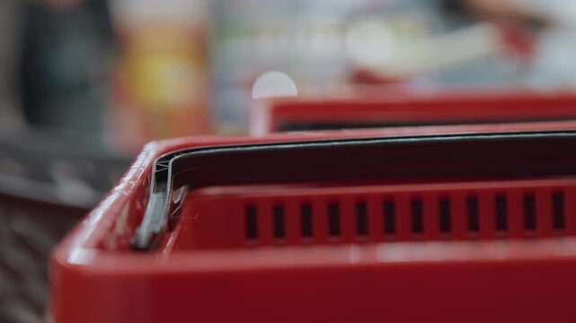 Red shopping basket handles in supermarket, closeup shallow depth of field showing worn black handle and plastic grid, blurred shoppers in background, shopper lifts basket by handle, muted fluorescent