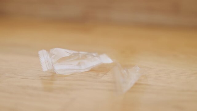 Small clear wrapper fragment resting on wooden table surface, crisp edges and soft reflection emphasize tiny debris and quiet aftermath of candy, intimate macro study of household litter.